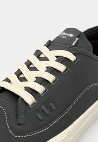 Mørk grå sneakers med en glatt tekstur, utstyrt med kremfargede snørebånd, hvite sømmer og tre teksturerte detaljer på siden.