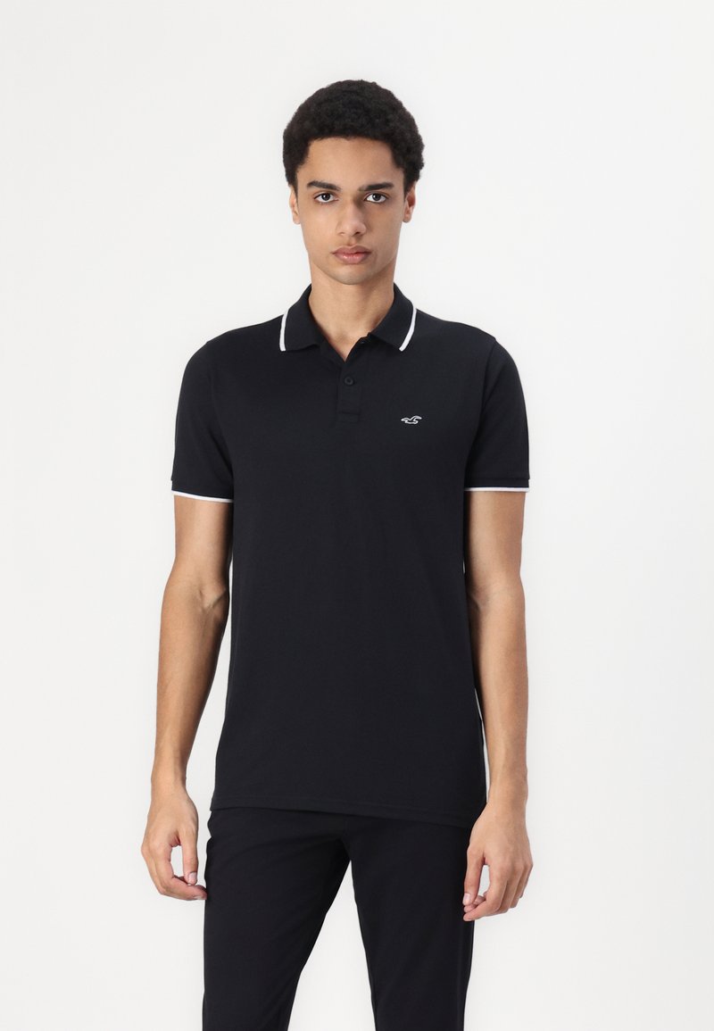 Polo noir avec un col et des poignets blancs contrastants, fabriqué dans un tissu lisse, avec un petit logo sur la poitrine.