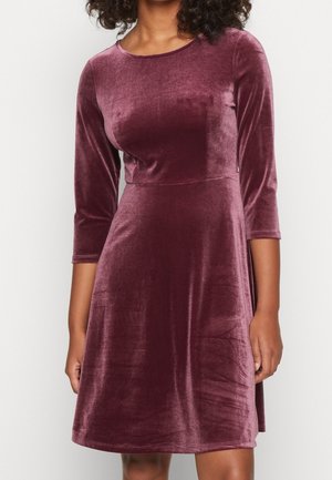 Femme portant une robe en velours prune, à manches longues, longueur genou, avec une taille cintrée et un col rond.