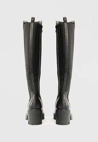 Botas negras hasta la rodilla con un exterior de cuero suave, paneles de tela texturizada, un tacón de bloque y un cierre de cremallera en la parte trasera.