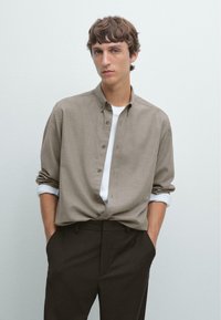 Young man with short brown hair wears a light brown button-up shirt over a white top, with dark pants and hands in pockets against a plain background.