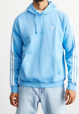 Mand iført en lyseblå Adidas hættetrøje med hvide striber på ærmerne og forlommer, kombineret med lyseblå jeans.
