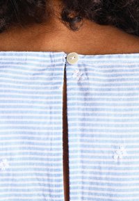 Tissu rayé bleu clair et blanc avec une fente arrière, fermée par un bouton unique. Présente de petites broderies près de la fente.