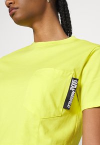 Klart gul bomullströja med rund halsringning, korta ärmar, en bröstficka och en svart etikett som accentuerar fickan.