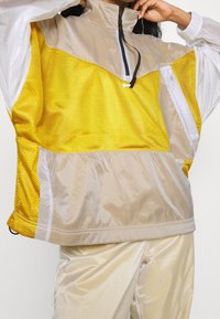Gul meshtröja med hög krage och dragkedja, med transparenta paneler och vita detaljer; strukturerad tyg med en lös passform.