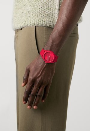 Bras à la peau foncée portant une montre-bracelet rouge avec un cadran à motif, reposant à côté d'un pantalon kaki et d'un pull texturé de couleur claire.