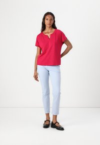 Helle pinke Bluse mit kurzen Ärmeln und V-Ausschnitt, kombiniert mit hellblauen Slim-Fit-Hosen und schwarzen Lackschuhen. Schlichtes, sauberes Design.