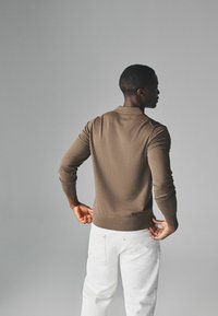 Brauner Strick-Polo-Pullover mit figurnaher Passform, gerippten Bündchen und Saum; kombiniert mit weißen Hosen. Modell von hinten gezeigt.
