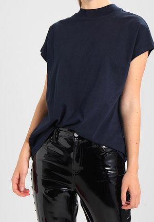 Personne portant un haut sans manches bleu marine rentré dans un pantalon en cuir verni noir brillant à taille haute sur un fond uni.