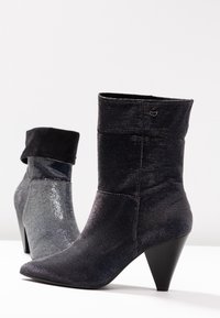 Bottes de cheville noires et argentées, avec un tissu texturé, un bout pointu et un talon conique. La botte noire possède un détail en cuir suédé au niveau du haut.