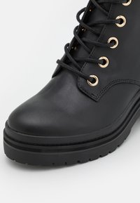 Botte noire en cuir avec lacets, œillets dorés et semelle en caoutchouc épaisse, montrant les détails de face et de côté.