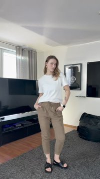 Wit T-shirt, bruine joggingbroek met trekkoord en zwarte sandalen. Kamer met een tv en een grijs tapijt op de achtergrond.