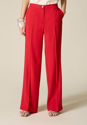 Kvinde iført højtaljede, vidde røde bukser med pressede folder, kombineret med en hvid top, klar armbånd og sandaler med slangeprint.