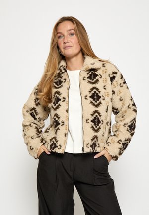 Tekoalaisa takki kermavärissä mustan ja ruskean geometrisen kuvion kanssa, vetoketjukiinnitys ja pehmeä rakenne. Sisältää pystykauluksen ja sivutaskut.