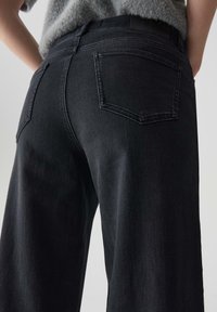 Jeans neri a gamba larga con una texture liscia, dotati di due tasche posteriori e una vita alta, mostrati da un'angolazione posteriore.
