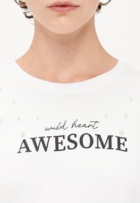 Weißes Baumwoll-T-Shirt mit der Aufschrift "wild heart" in Schreibschrift und "AWESOME" in fettem schwarzen Text, verziert mit kleinen, perlenähnlichen Verzierungen.