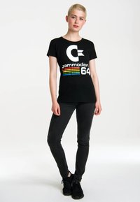 Black t-shirt featuring a large white "C" logo and "commodore 64" text with colorful stripes underneath, paired with dark jeans and black sneakers.