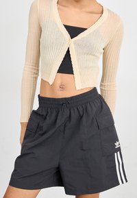 Béžový žebrovaný kardigan s vroubkovanými okraji, nošený přes černý top. Černé šortky s bočními kapsami a bílými pruhy; zobrazen logo Adidas.