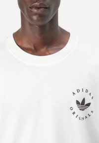 Balta medvilninė marškinė su apvaliu iškirpimu, trumpomis rankovėmis ir juodu „Adidas Originals“ logotipu, atspaustu ant krūtinės.