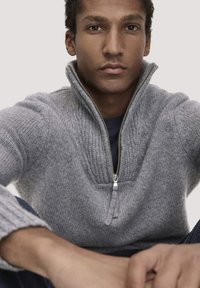 Grau gestrickter Halb-Reißverschluss-Pullover mit strukturierten Mustern und gerippten Bündchen. Verfügt über einen silbernen Reißverschluss und eine lockere Passform, die über einem dunklen Oberteil getragen wird.