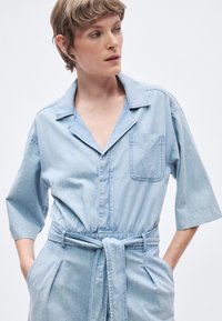 Φως μπλε τζιν φορμάκι με γιακά στο λαιμό, κοντά μανίκια και δεμένη μέση. Διαθέτει τσέπη στο στήθος και πτυχώσεις στο μπροστινό μέρος.