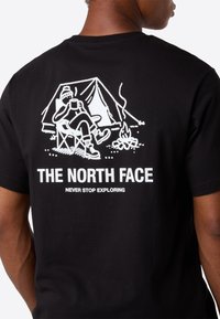 Bagsiden af en sort t-shirt med hvid grafik af en person, der drikker ved et lejrbål foran et telt, med teksten "THE NORTH FACE NEVER STOP EXPLORING."