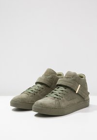 Zapatillas altas verdes de ante con un acabado texturizado, que presentan un cordón en la parte frontal y una correa alrededor del tobillo con un acento metálico.