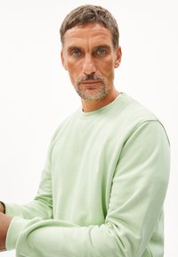 Sweatshirt vert clair en matériau doux avec un col rond et des manches longues ; présente un petit logo blanc sur la poitrine.
