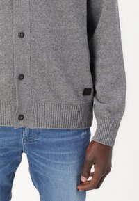 Cardigan de punto gris con dobladillo acanalado, botones negros en la parte delantera y una pequeña etiqueta negra en el lado. Lleva puesto sobre jeans azules.