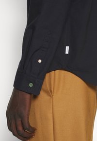 Gros plan sur une main à la peau foncée à côté d'un pantalon couleur moutarde, portant une chemise noire à manches longues avec des boutons dépareillés au niveau du poignet.