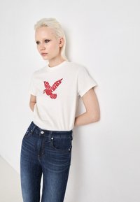 Weißes T-Shirt mit einem roten Vogelmotiv in der Mitte, kombiniert mit dunkelblauen Jeans. Aus Baumwolle mit einer lockeren Passform.