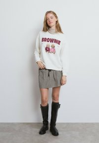 Sudadera blanca con el gráfico "BROWNIE", combinada con una falda a cuadros marrón y beige. La modelo lleva botas negras altas, de pie frente a un fondo liso.