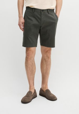 Man draagt donkergroene knielange shorts, beige overhemd en bruine instapschoenen, staat met handen in de zakken tegen een effen achtergrond.