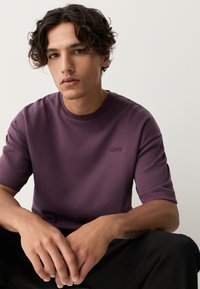 Lila T-Shirt mit kurzen Ärmeln aus weichem Stoff, mit rundem Ausschnitt und kleinem Logo auf der Brust. Glatte, gleichmäßige Textur.