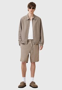 Ljusbrun, lättviktig jacka med krage, i kombination med beiga shorts. Modellen bär solglasögon och bruna skor, med vita strumpor.