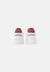 Zapatillas blancas con una textura suave, que presentan una lengüeta trasera roja etiquetada con "DESDE 1890" y un sutil cosido blanco. Diseño de perfil bajo.