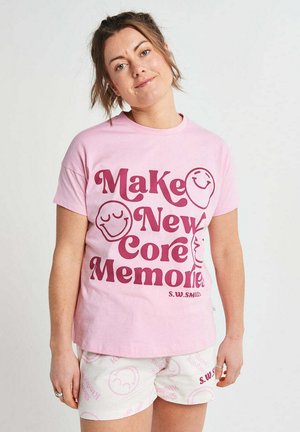 Frau trägt ein pinkes T-Shirt mit dem Schriftzug „Make New Core Memories“ und lächelnden Gesichtern, kombiniert mit weißen Shorts mit pinkem Smiley-Muster.