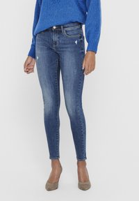 Jean skinny bleu foncé en denim, avec des accents usés et des décolorations, assorti à des chaussures à talons hautes beige et un pull bleu.