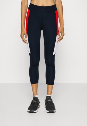 Femme portant des leggings bleu marine courts avec des panneaux latéraux rouges et blancs, assortis à des baskets noires et des chaussettes blanches, debout devant un fond blanc.
