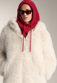 Plašč iz umetnega krzna v kremni barvi, z ruto in zadrgo. Kombiniran z rdečo pleteno ruto in temnimi sončnimi očali. Mehka tekstura, prevelika oblika.