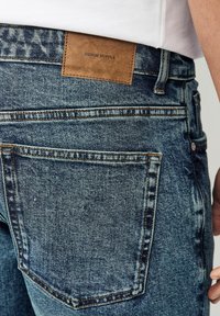 Jeans din denim de culoare albastră închisă cu un finisaj uzat, având o etichetă din piele cu inscripția "DENIM SUPPLY" și buzunare standard pe spate.