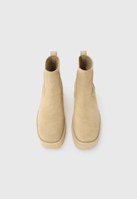 Et par beige suede ankelstøvler med runde firkantede tæer og trækflapper, vist ovenfra på en hvid baggrund.