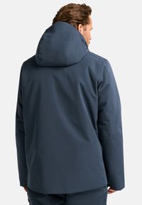 Giacca impermeabile blu navy con un ampio cappuccio, texture morbida e vestibilità rilassata. I dettagli delle cuciture sono visibili sul retro.