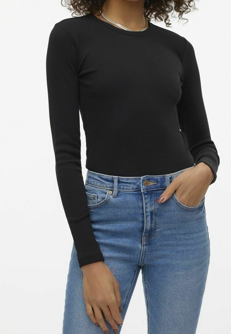 Frau in einer figurbetonten schwarzen Bluse mit langen Ärmeln und hoch taillierten hellblauen Jeans, mit einer Hand in der Tasche, vor einem weißen Hintergrund.