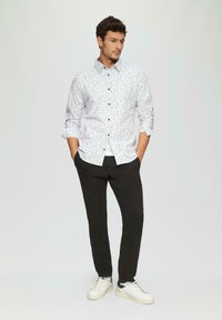 Witte button-up blouse met een blauw abstract patroon, gecombineerd met zwarte broek en witte sneakers, met een relaxte fitting en opgerolde mouwen.