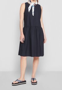 Vestido sin mangas de color navy con cintura fruncida, textura suave, combinado con chanclas a rayas azul marino y blancas, y una bufanda blanca atada al cuello.