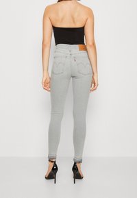 Mujer vistiendo jeans ajustados de tiro alto en gris claro con dobladillos deshilachados y zapatos de tacón negro con tiras, mostrada desde atrás.