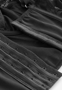 Corset negru cu închideri cu cârlige și ochiuri, având o textură netedă și accente de dantelă intricate. Straturi de țesătură creează structură și suport.