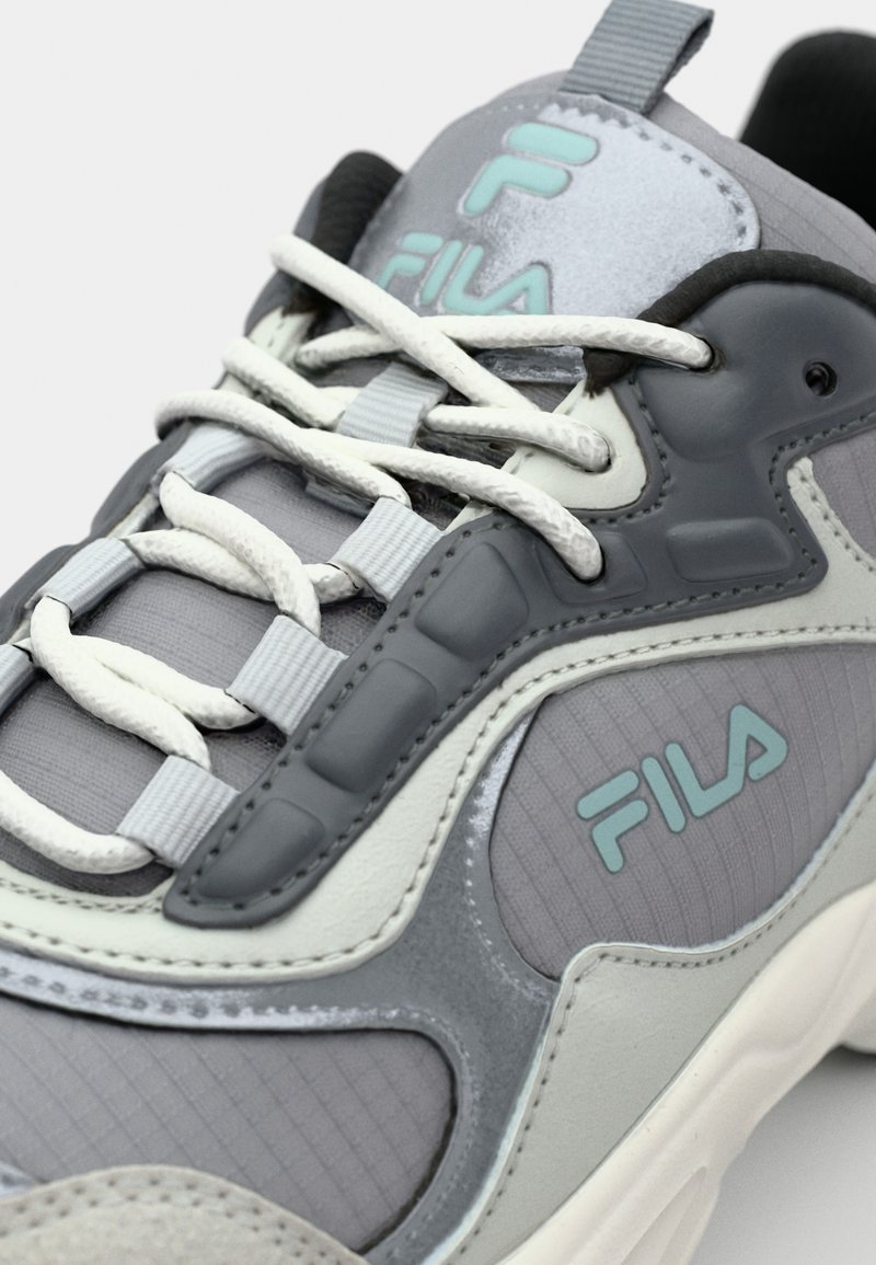 Sneaker grise présentant un mélange de tissus texturés et de cuir lisse. Des accents bleu clair et un logo bien en vue sur le côté rehaussent le design.