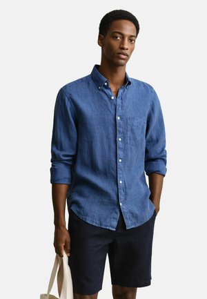 Man wearing a blue button-up shirt and black shorts, holding a beige tote bag, standing with one hand in pocket against white background.